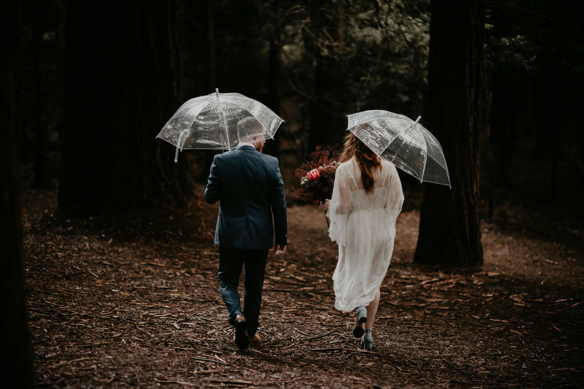 bride and groom walk through trees with umbrella on a rainy day in the Warburton Redwood Forest after their wedding with Let's Elope Melbourne Celebrant beautiful ceremony and photography Elopement Packages Victoria Sarah Matler Photography