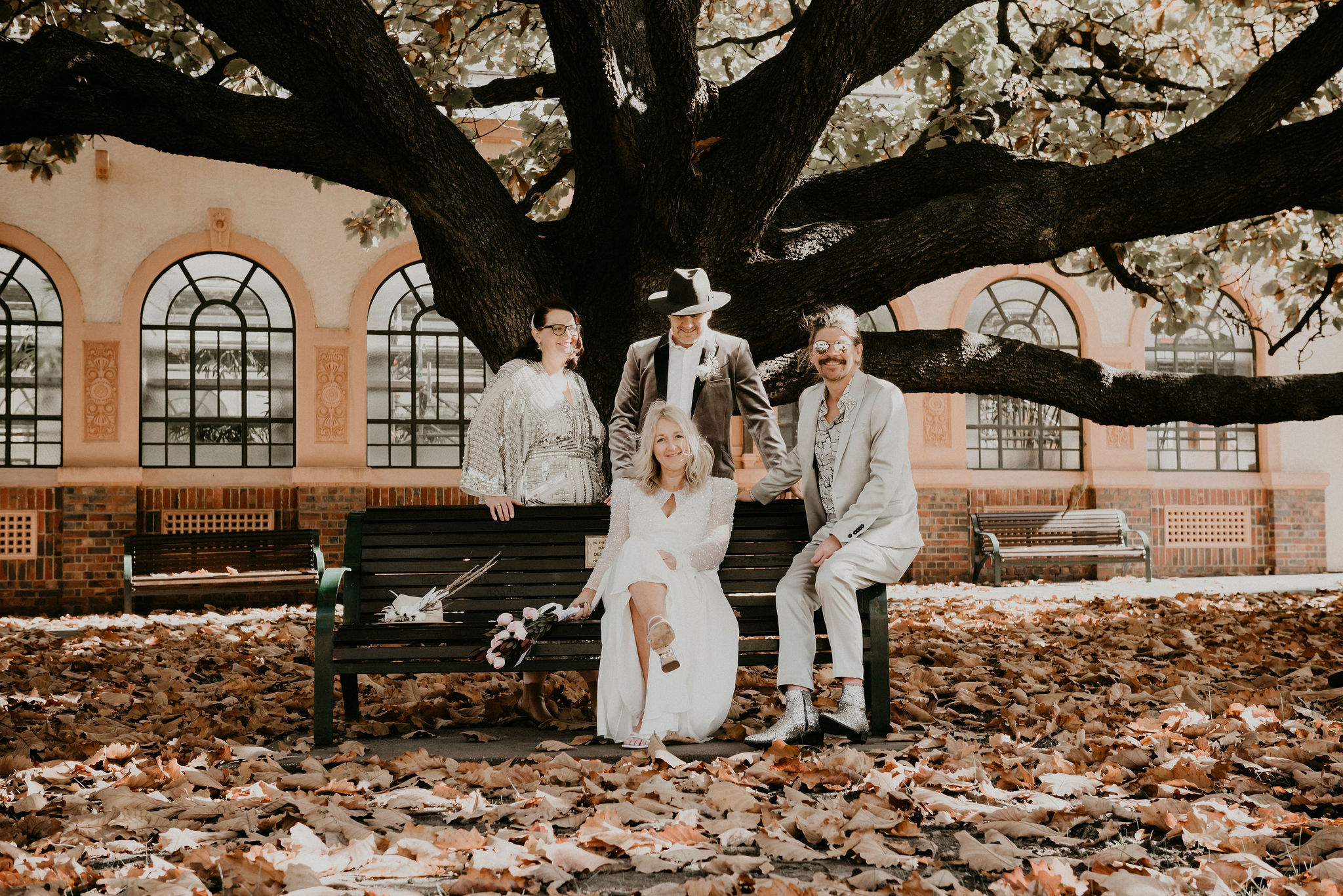 bride groom couple and witness sit under autumn tree with surrounded by leaves wearing sequins hats silver boots velvet jacket Fitzroy Gardens Melbourne Laneways Elopement Lets Elope Melbourne Celebrant ceremony and photography Elopement Packages Victoria Sarah Matler Photography Fitzroy Gardens City Wedding Photos ACDC Lane Laneways Alternative