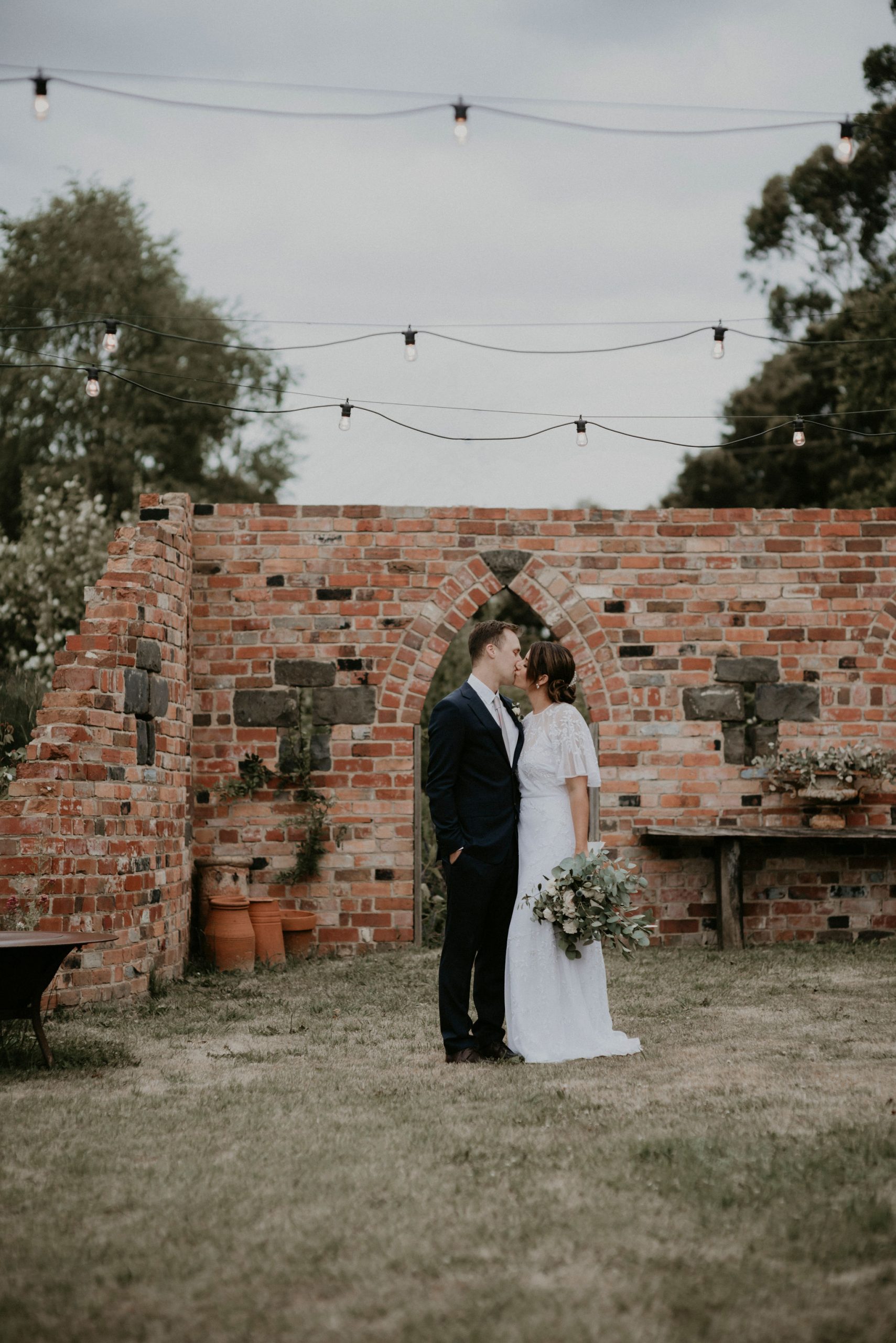 Couple kiss in front of brick wall arch with fairy lights Let's Elope Melbourne Portfolio Celebrant Photographer Elopement Packages Victoria Sarah Matler Photography intimate weddings Acre of Roses Trentham romantic ceremony