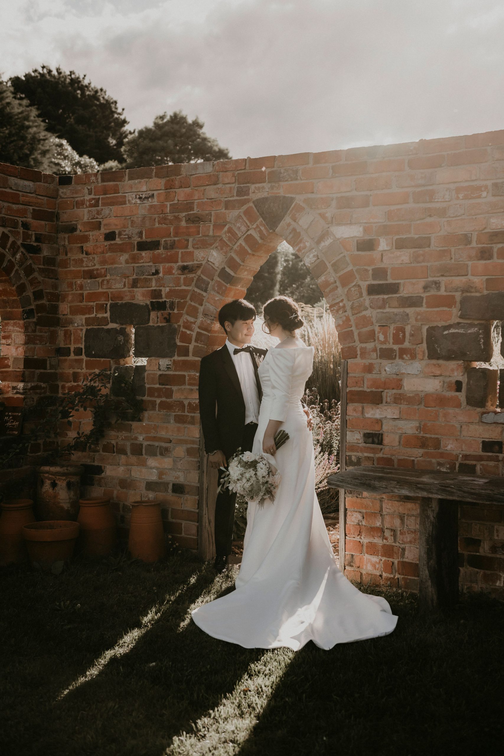 Bride and groom stand closely by the brick folly bathed in afternoon sunlight with close family Let's Elope Melbourne Portfolio Celebrant Photographer Elopement Packages Victoria Sarah Matler Photography intimate weddings Acre of Roses Trentham romantic ceremony