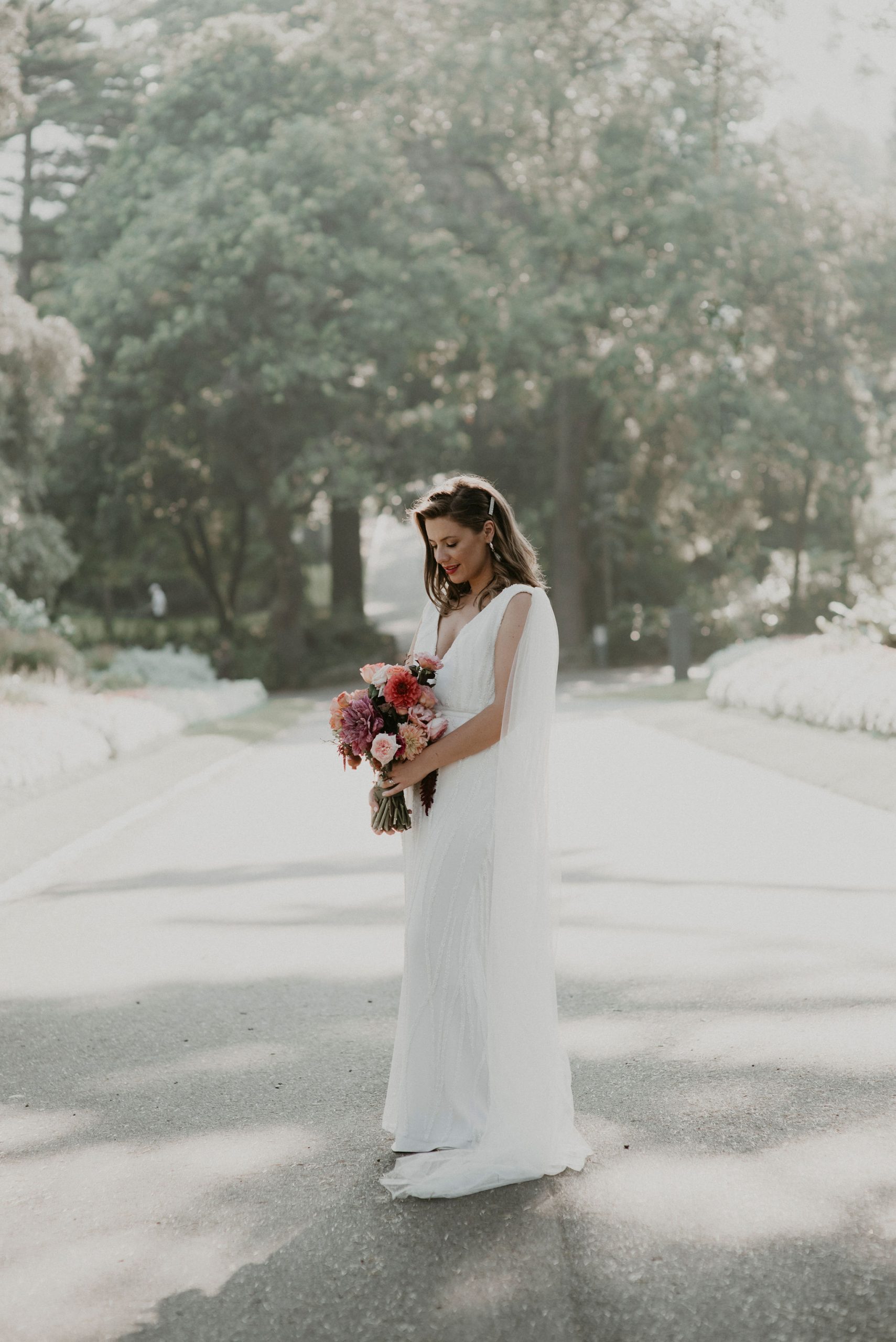 Bride stands in the middle of path looking down at flowers bathed in sunlight Let's Elope Melbourne Portfolio Celebrant Photographer Elopement Packages Victoria Sarah Matler Photography intimate wedding Fitzroy Gardens East Melbourne elopement