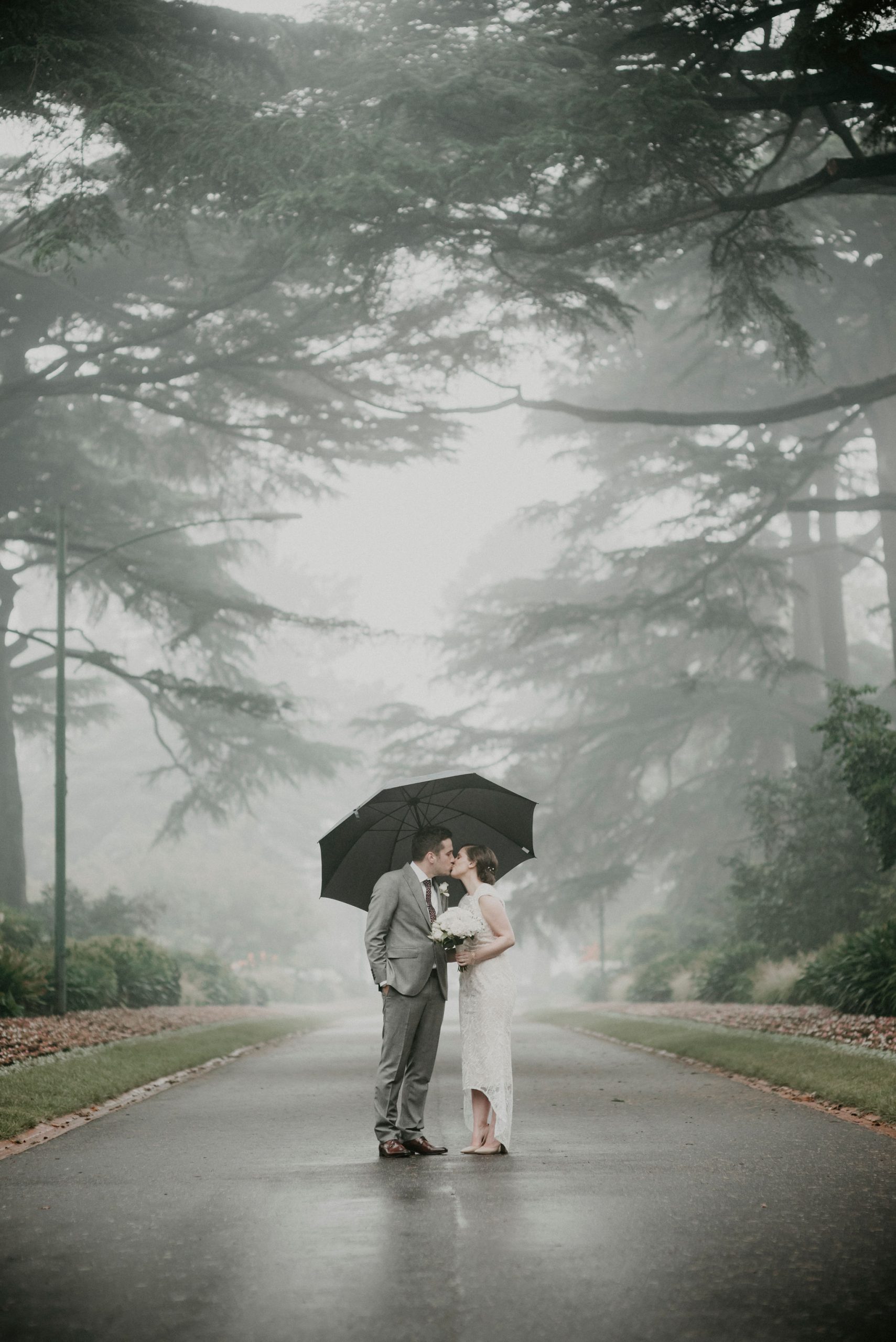 Bride and groom kiss in the middle of the pathway in the rain and fog holding an umbrella Let's Elope Melbourne Portfolio Celebrant Photographer Elopement Package Victoria Sarah Matler Photography intimate wedding Fitzroy Gardens Fog Moody East Melbourne